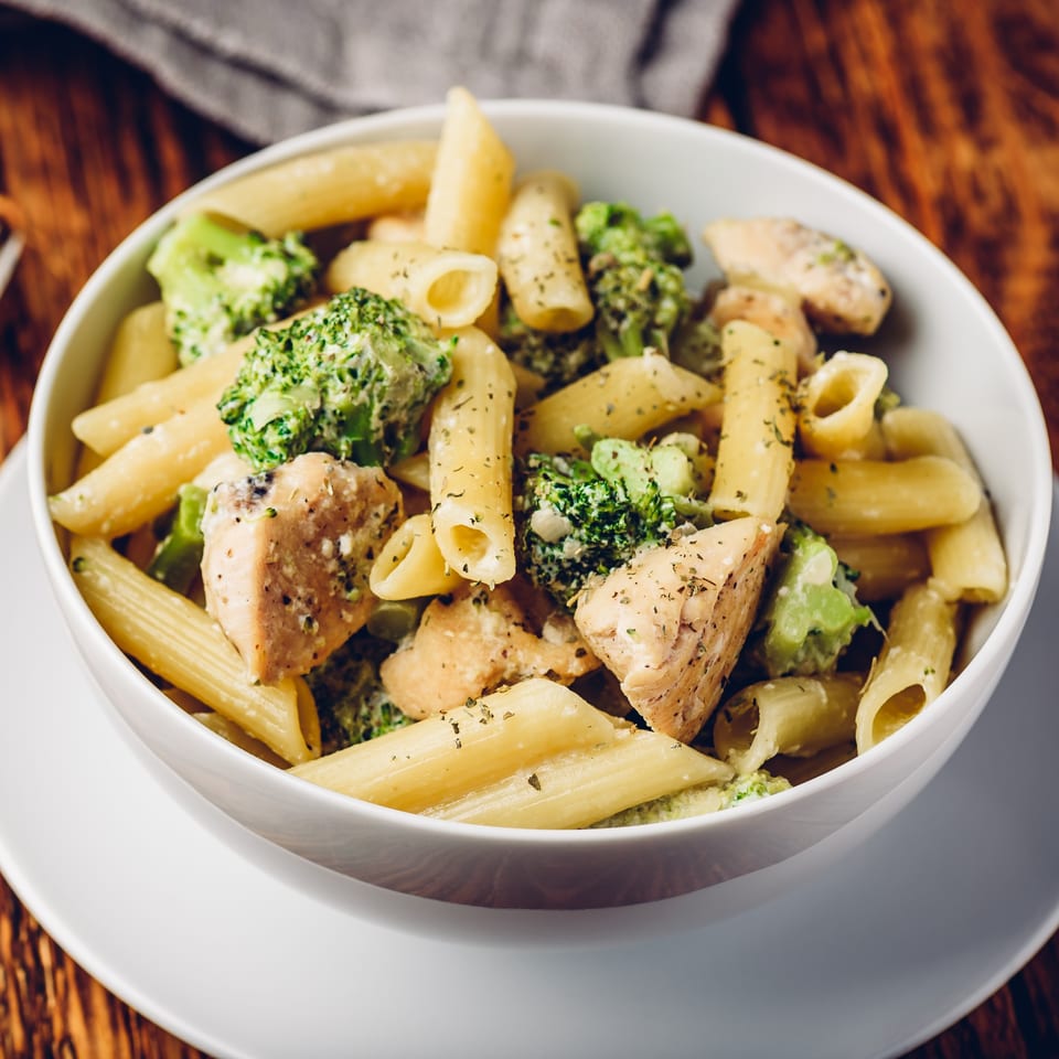 Penne pasta with chicken, broccoli, and creamy sauce, garnished with herbs in a white bowl.