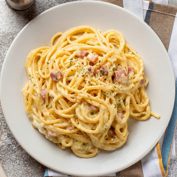 Creamy spaghetti carbonara with bacon bits and parsley on a white plate, next to a striped napkin.